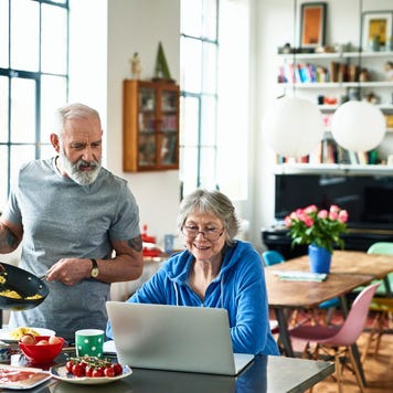 Senior woman using laptop and smiling as man serves breakfast