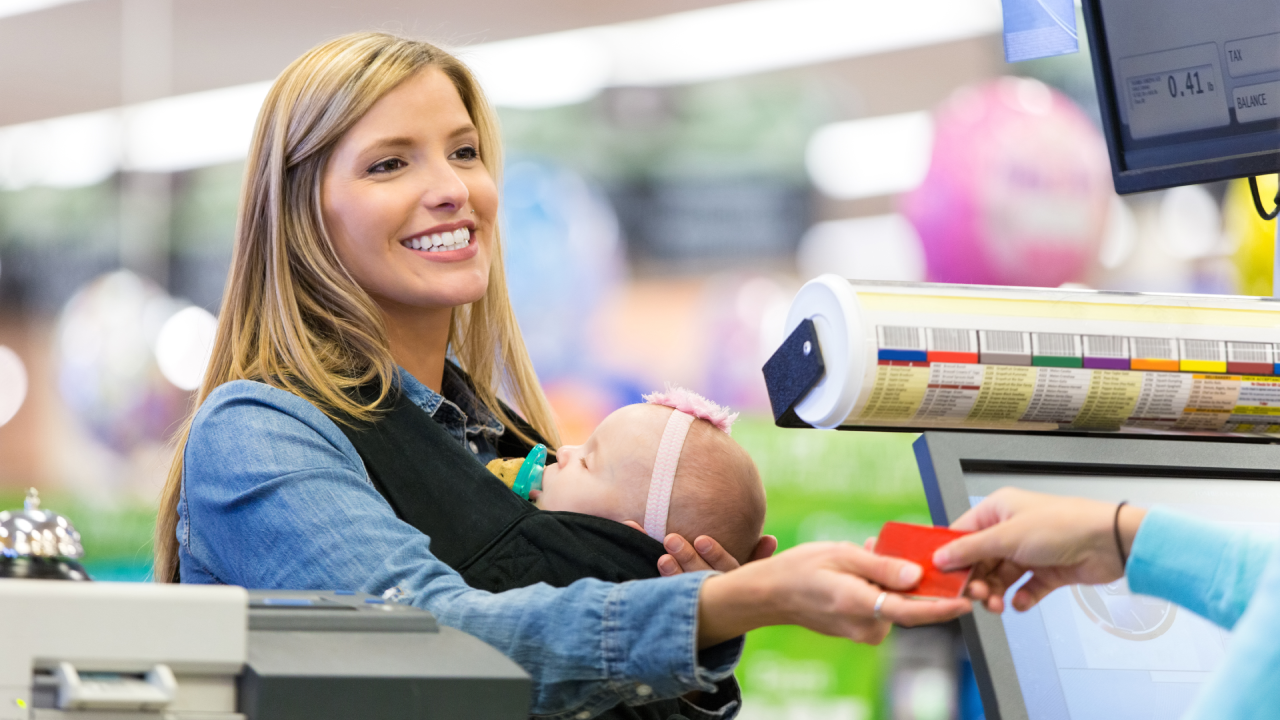 Mid adult Caucasian woman is smiling while shopping in grocery store with infant daughter. Woman is handing loyalty card or credit card to grocery store employee cashier. She is wearing her baby in a baby wearing wrap carrier. Customer is purchasing food and other items.