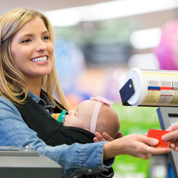 Mid adult Caucasian woman is smiling while shopping in grocery store with infant daughter. Woman is handing loyalty card or credit card to grocery store employee cashier. She is wearing her baby in a baby wearing wrap carrier. Customer is purchasing food and other items.