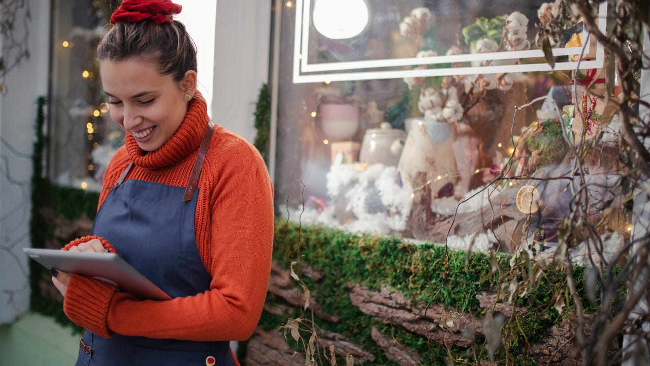 A smiling store owner in a turtleneck sweater stands outside her shop, looking at her tablet.