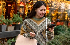 A young smiling woman stands outside of a festive store, looking at her phone.