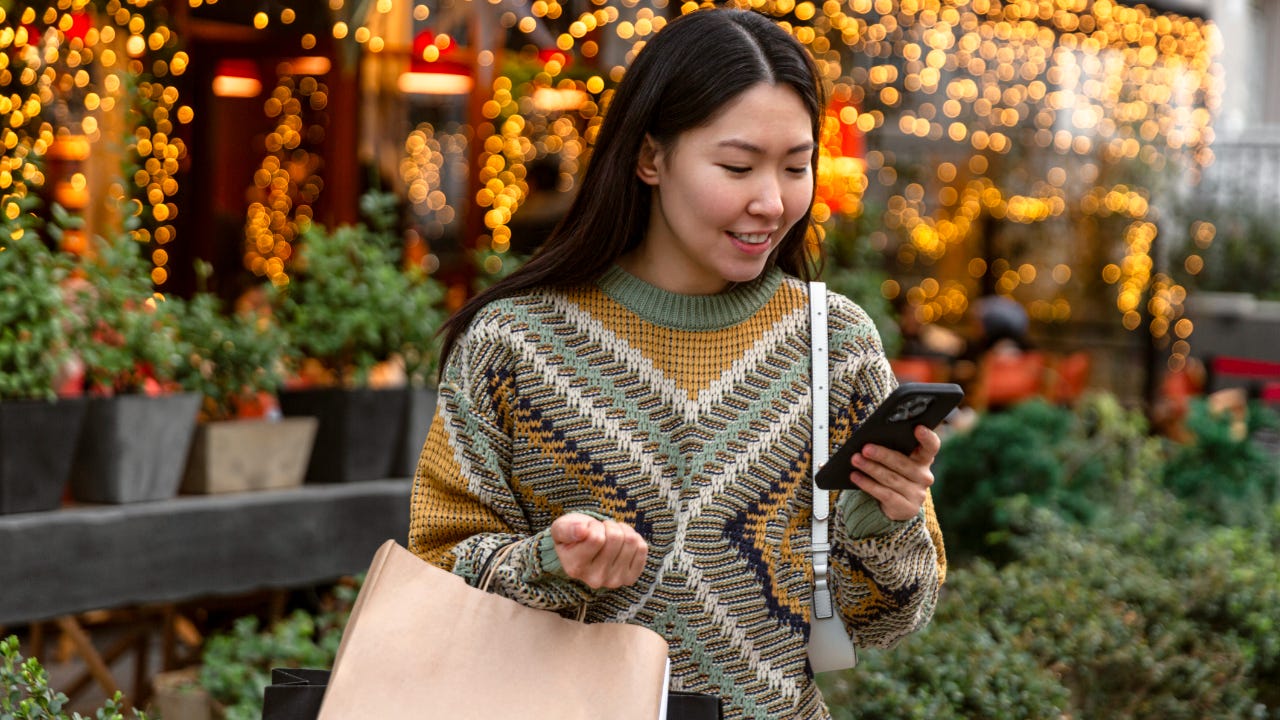 A young smiling woman stands outside of a festive store, looking at her phone.