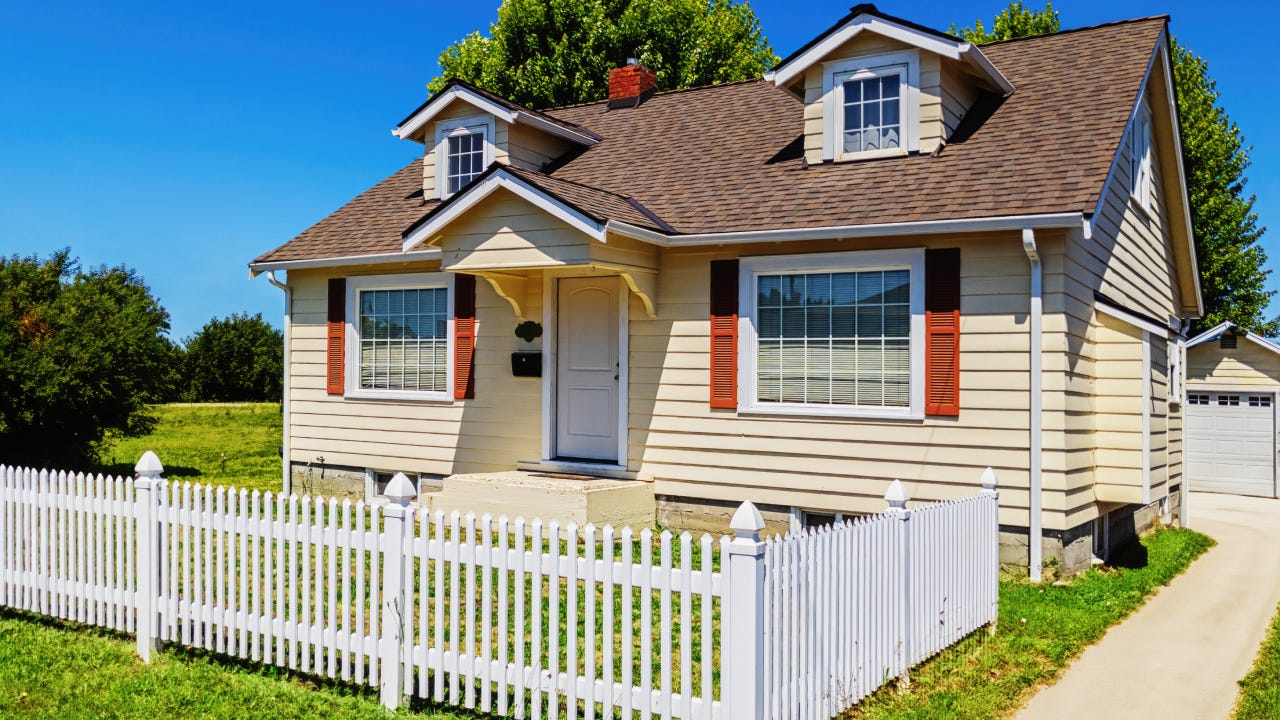 Photo of a small yellow suburban home with a white picket fence surrounding the front yard, on a sunny summer day