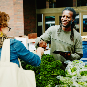 Smiling organic farmer taking credit card from customer to run transaction on digital tablet at farmers market