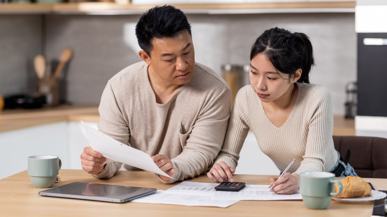 Serious asian family husband and wife sitting at kitchen table full of papers, laptop, coffee mugs, counting monthly expenses, using calculator, short of money. Economy, family budget concept