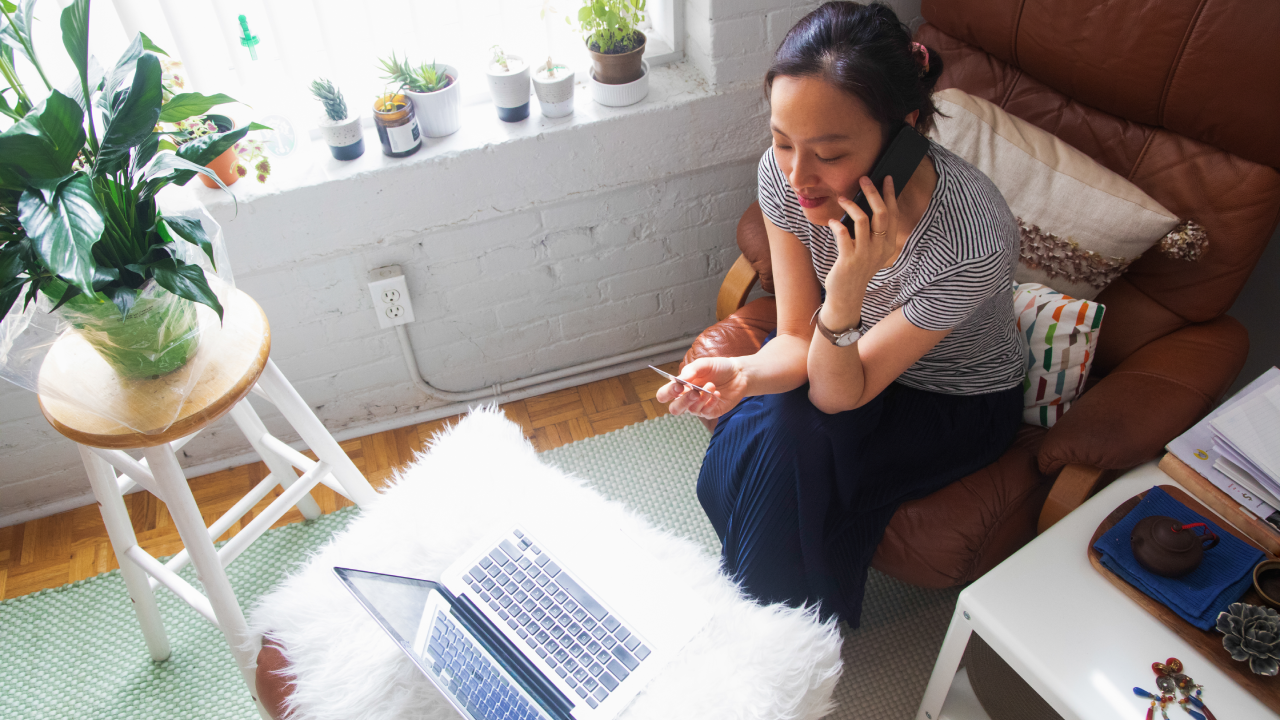 Overhead view of young Asian woman talking to customer service, credit card in hand, in her living room with a laptop in front of her.