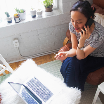 Overhead view of young Asian woman talking to customer service, credit card in hand, in her living room with a laptop in front of her.