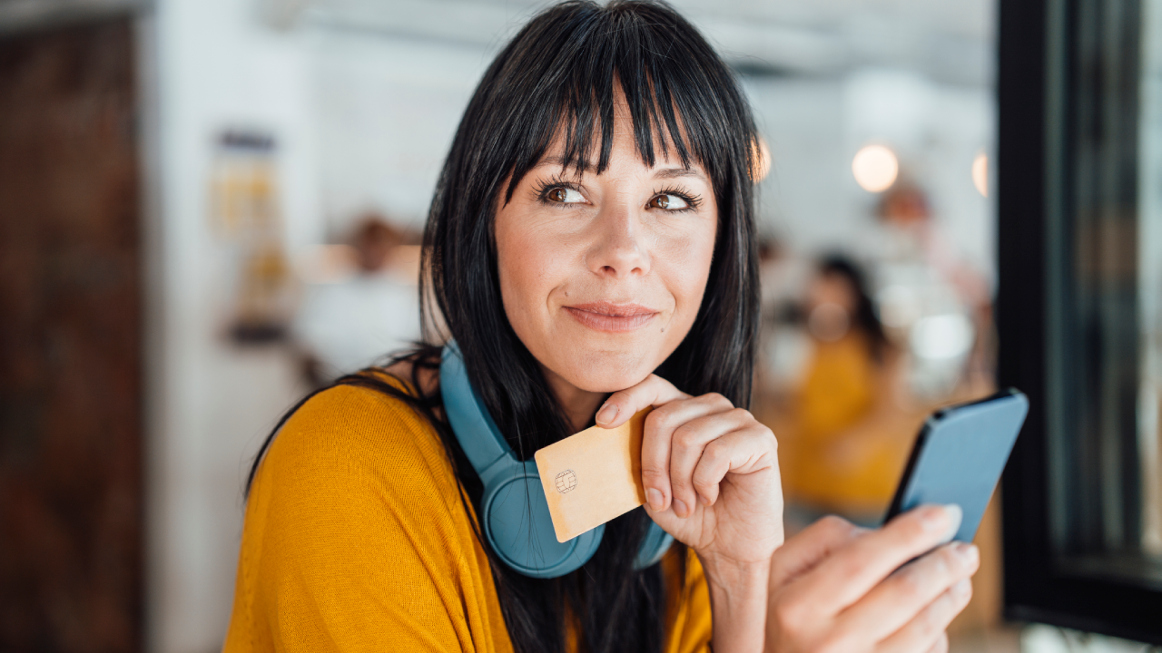 Contemplative woman with with headphones around neck holding credit card and smart phone - stock photo
