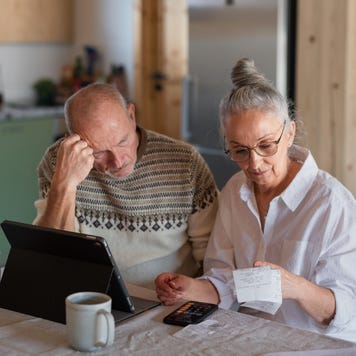 Senior couple sitting at the kitchen table with croissant and cups of tea, looking at digital tablet and recalculating their expenses.
