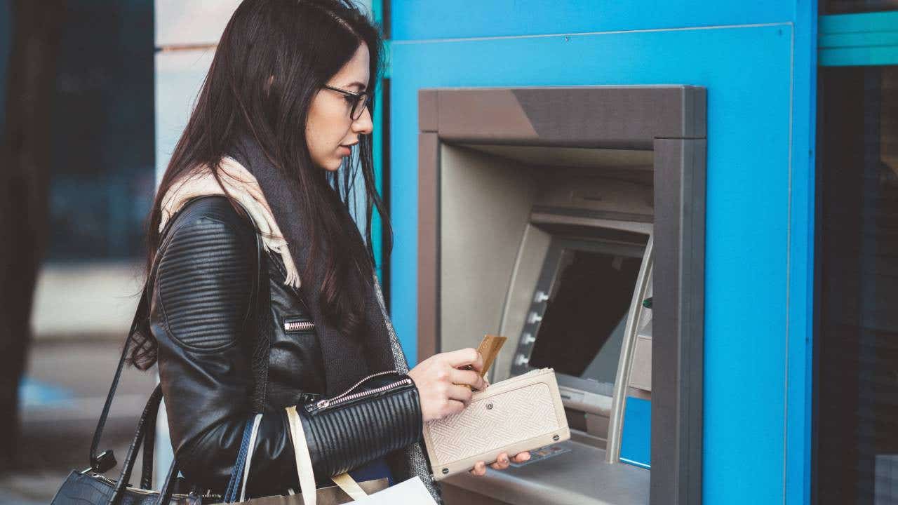 A person in casual attire stands at an ATM machine, inserting a card and interacting with the keypad to carry out a transaction.