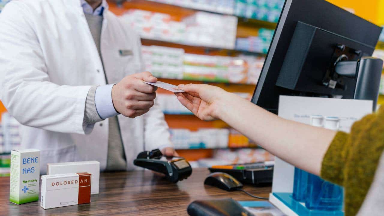 A person with outstretched hand handing a credit card to a pharmacist for payment. The pharmacist stands behind a counter, ready to process the transaction.