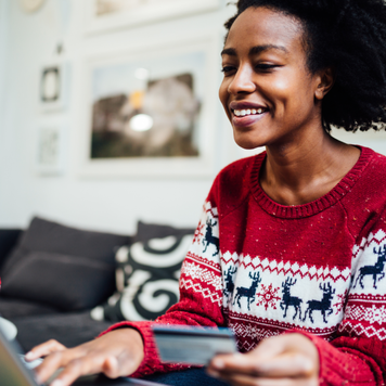 Beautiful African American woman shopping online from her cozy home office during winter holidays, holding a credit card while purchasing something online using a laptop