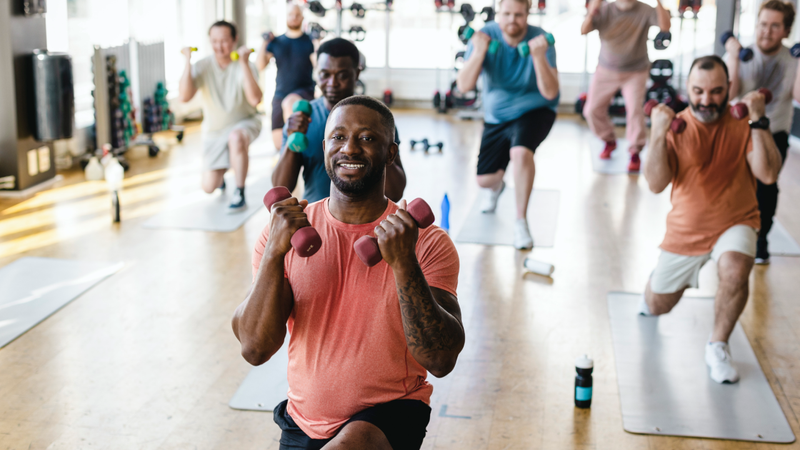 Smiling male fitness instructor practicing dumbbell exercise with men in gym - stock photo
