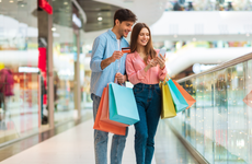 Shopping Application. Happy Husband And Wife Using Smartphone And Credit Card Holding Colorful Shopper Bags And Using Mobile App Buying Clothes Online In Modern Mall Indoor. Ecommerce Concept