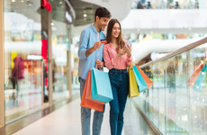 Shopping Application. Happy Husband And Wife Using Smartphone And Credit Card Holding Colorful Shopper Bags And Using Mobile App Buying Clothes Online In Modern Mall Indoor. Ecommerce Concept