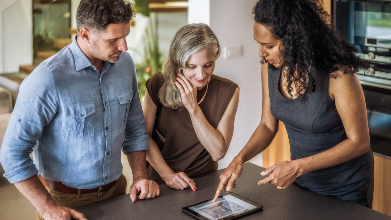 Real estate agent showing property for sale catalog on digital tablet to clients couple at kitchen counter