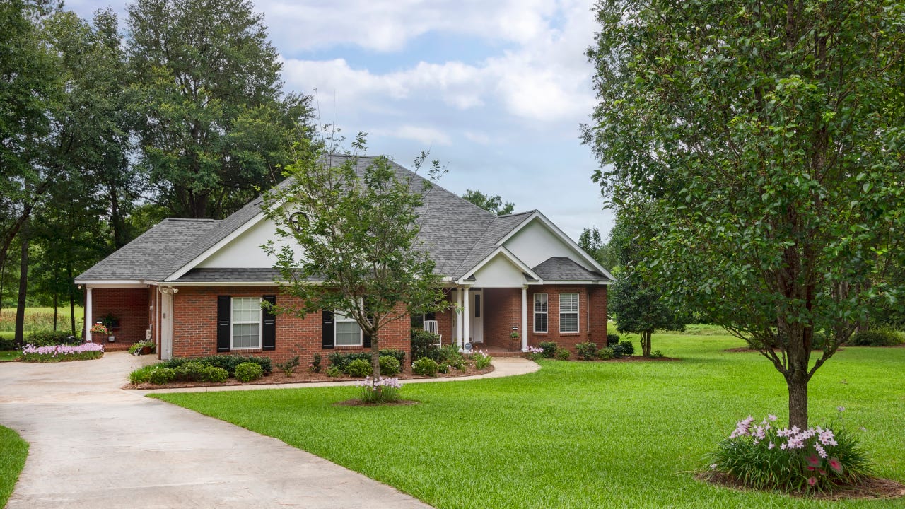 Traditional style brick house with black shutters, situated on a large green yard with mature trees.