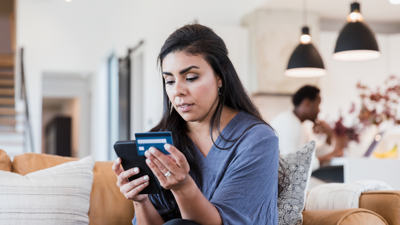 A serious mid adult woman enters her credit card number while shopping online. Her husband is working in the background.