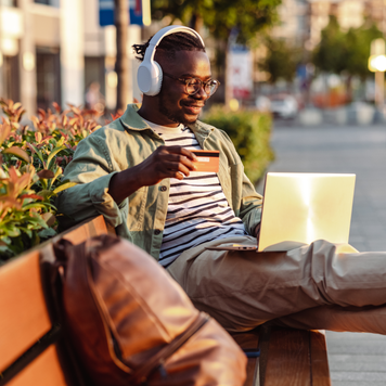 Photo of a smiling African-American man sitting on the bench and holding a credit card. E-commerce activity