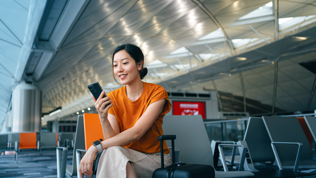 Young Asian woman with suitcase using smartphone while waiting for her flight at airport terminal. Asian businesswoman on business travel. Lifestyle and technology. Travel and vacation concept
