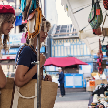 Two women are examining items for sale at an outdoor market in Etretat, Normandy, France. The young woman on the left is wearing a red beret, a red and white striped top. The mother, mature woman on the right, dressed in a navy-blue shirt and carrying a large straw bag.