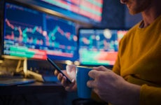 Man using mobile phone and having coffee while monitoring stock market on computer screens at desk