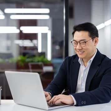 Asian man in business suit and glasses working with laptop inside office, mature businessman typing on keyboard, preparing financial report, male company employee at workplace.