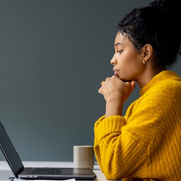 Black woman in an orange shirt viewed in profile looking down at her laptop thoughtfully with her chin on her hands.