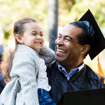 Older graduate smiling at his laughing granddaughter who he is holding
