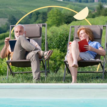 An older white couple sits together outside while reading books.