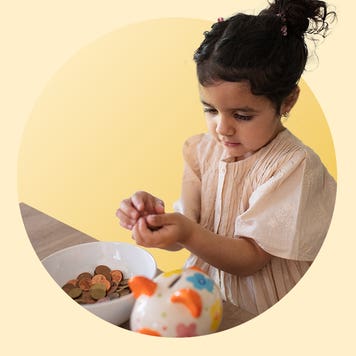 Image of a little girl holding coins in her hand. She sits in front of a table with a bowl full of coins and a multi-colored piggy bank.