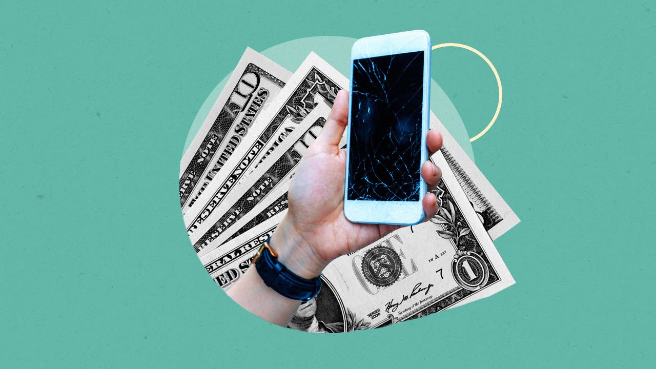 Close up of a hand holding a smartphone with a cracked screen against a background of dollar bills