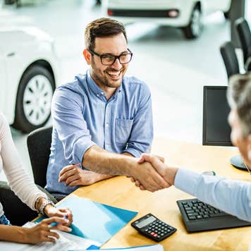 man and woman at a car dealership shaking hands with a salesperson after buying a car