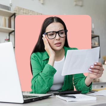 young woman in glasses sitting by her laptop reading a piece of paper in dismay