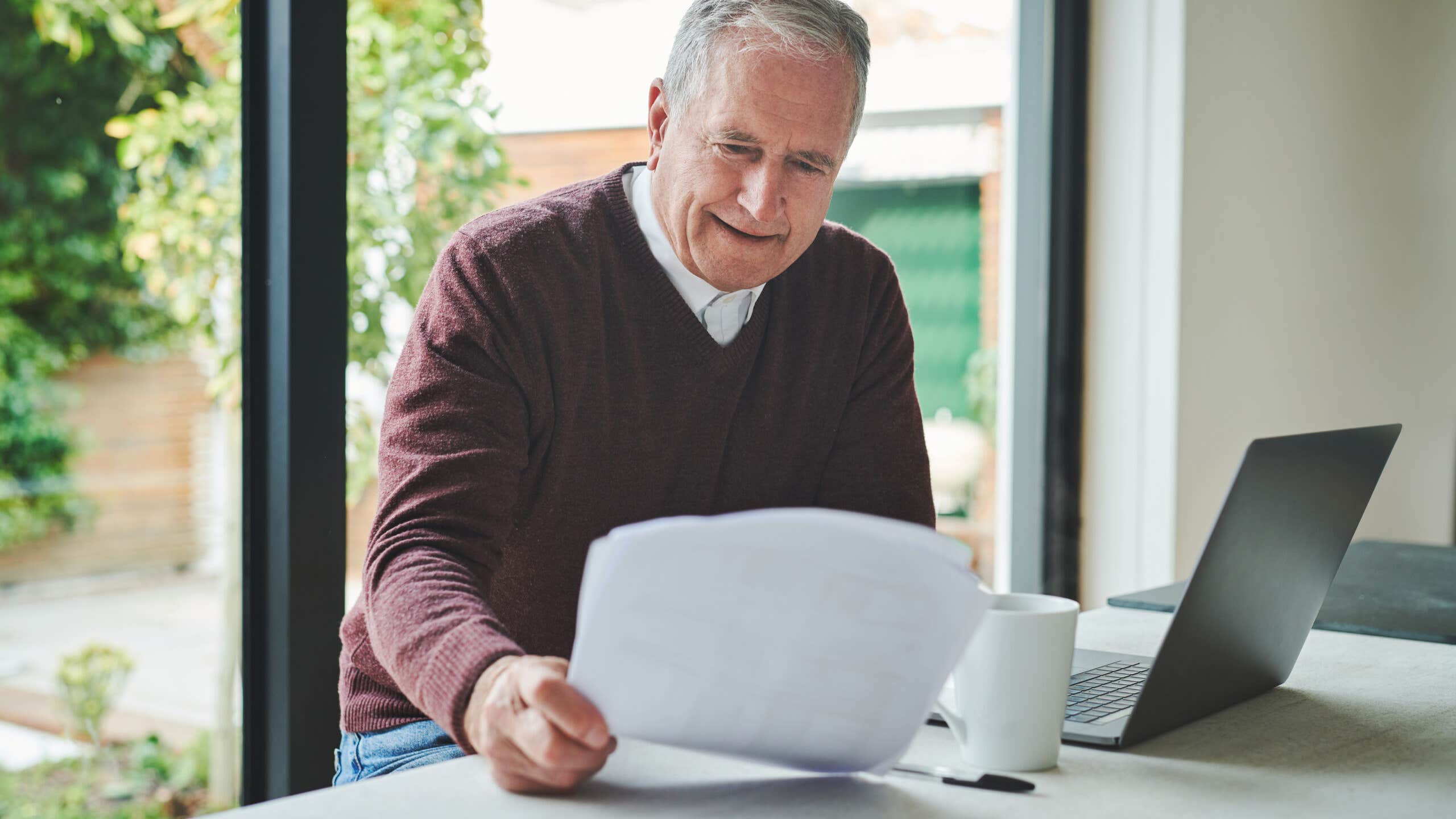 Elderly man looking at his insurance policy sitting next to an open laptop