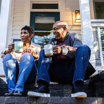 A couple sits on a porch of a house drinking coffee.