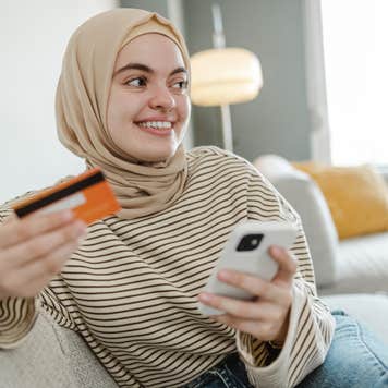 Photo of a smiling young woman holding a credit card and using a smart phone for online shopping