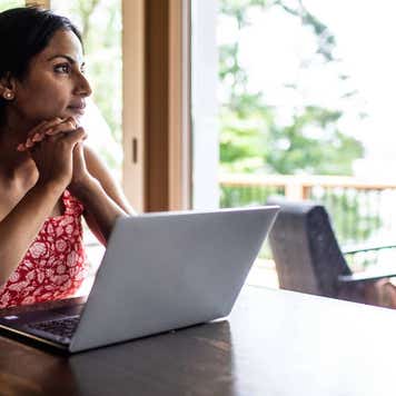 Middle aged woman looks out window sitting at table with laptop in front of her.