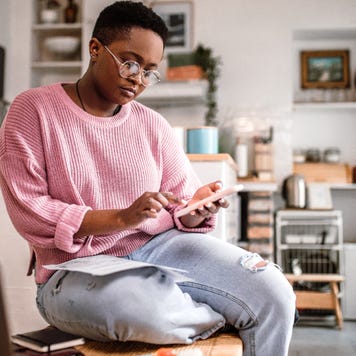 Black woman sitting in her apartment using her phone with her laptop in front of her.