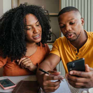 A young African-American man and woman sit at a table working on their finances with a laptop, phone and notebooks.
