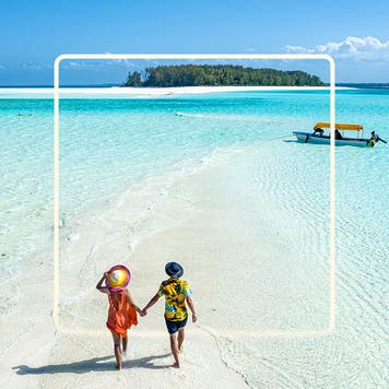 Image of two people in wide brim hats and summer clothes walking hand-in-hand down a narrow strip of white sand beach towards a small motor boat in the distance. Blue water laps in small waves on either side of the strip of beach.
