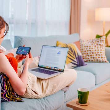 A young woman sits on her couch and trades stocks on her iPhone and computer.