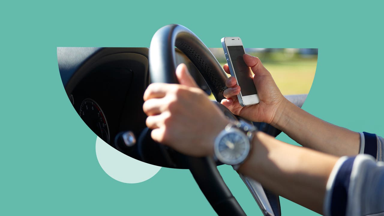 Close-up of hands on a steering wheel; one hand is holding a smartphone.