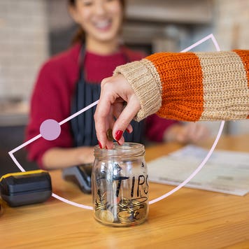 Image of a tip jar, filled with money, sitting on a counter. There is a hand adding more money to it while a smiling worker in an apron looks on.