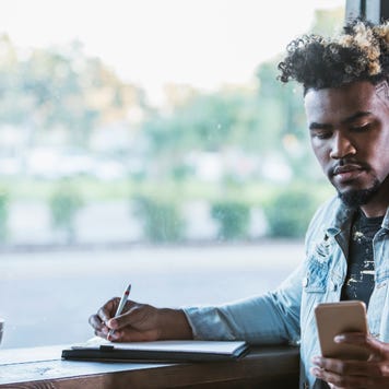Young Black man sitting at coffee shop and checking his phone while writing on a piece of paper.