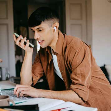 Young male freelancer talking on speaker phone while working on laptop at home office