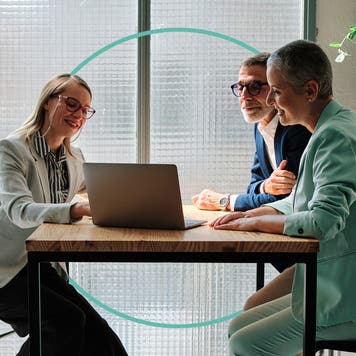 Two women and a man sit at a table and look at a laptop.