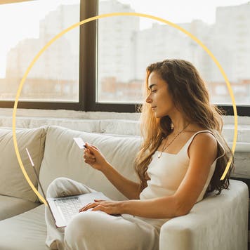 A young woman sits on a white couch looking at her credit card with a laptop on her lap. There is an illustrated yellow circle behind her.