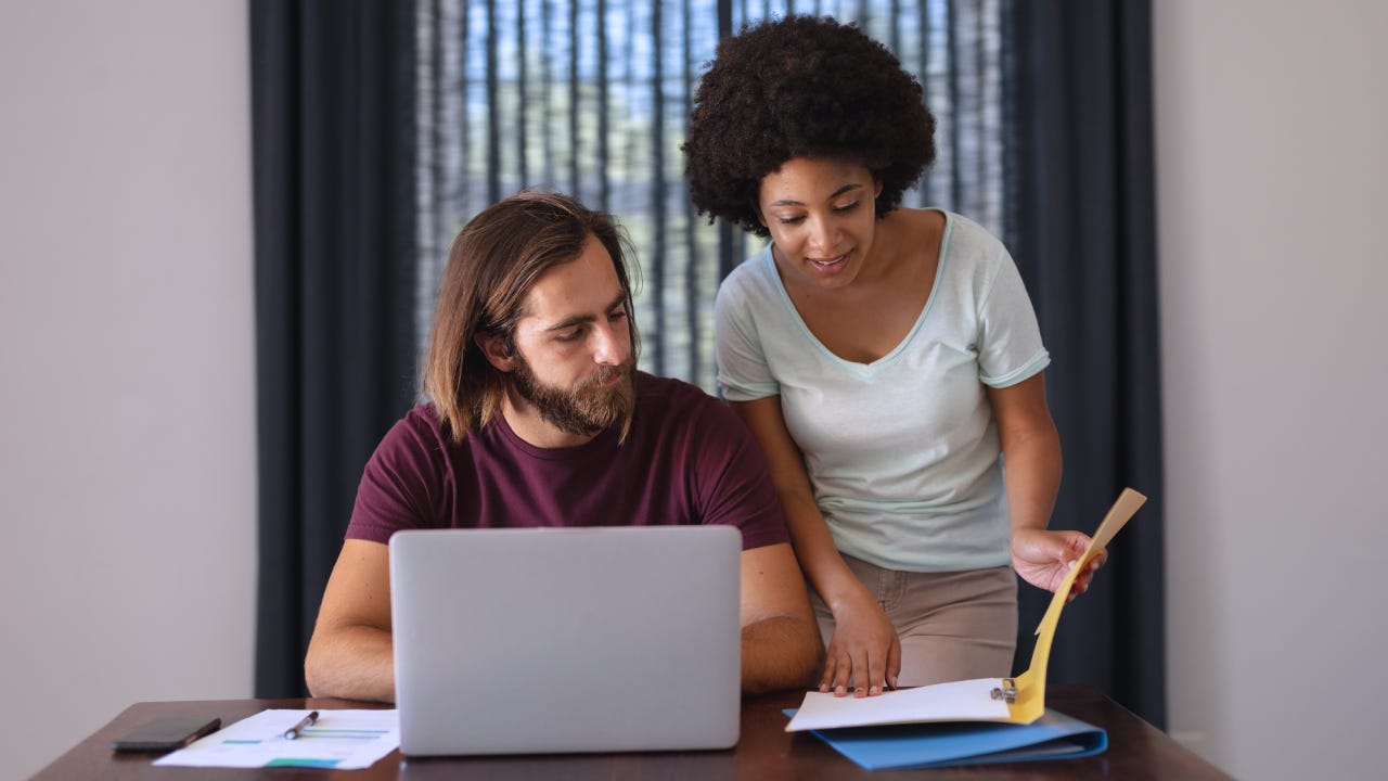A Black woman and a white man work together. The woman is standing and reviewing a book while the man is on a laptop.