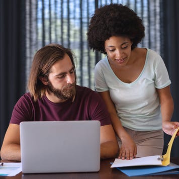 A Black woman and a white man work together. The woman is standing and reviewing a book while the man is on a laptop.